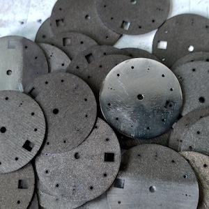 May include: A collection of gray watch dials with various hole sizes and square cutouts. The dials are circular, with a brushed metal finish on some and a textured, matte finish on others. The image shows a close-up view of the watch components.