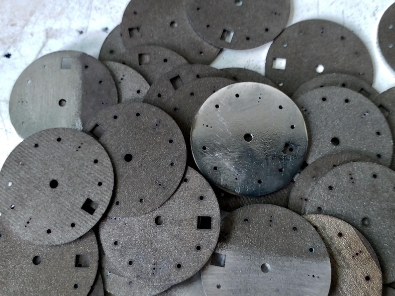 May include: A collection of gray watch dials with various hole sizes and square cutouts. The dials are circular, with a brushed metal finish on some and a textured, matte finish on others. The image shows a close-up view of the watch components.