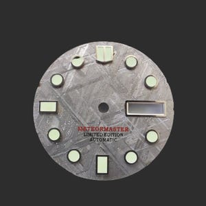 Puede incluir: Una esfera de reloj circular con una superficie gris texturizada. Cuenta con marcadores y agujas luminosas, con el texto "METEORMASTER LIMITED EDITION AUTOMATIC" en rojo. El diseño incluye un agujero central y una ventana rectangular.