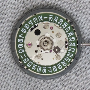 May include: Close-up of a watch movement with a green date dial and silver gears. The dial displays numbers 1-31 in white. The watch mechanism is set against a grey background.