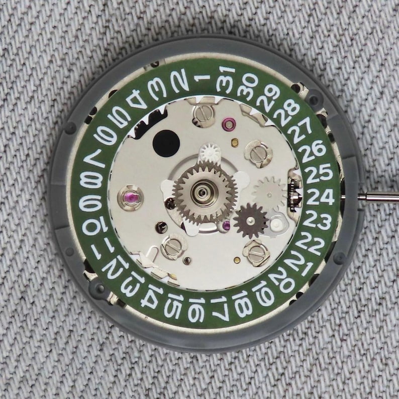 May include: Close-up of a watch movement with a green date dial and silver gears. The dial displays numbers 1-31 in white. The watch mechanism is set against a grey background.