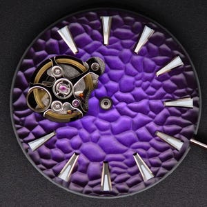 Puede incluir: Primer plano de una esfera de reloj con un fondo morado intenso texturizado. Los marcadores de hora plateados y blancos están dispuestos alrededor del perímetro. El mecanismo del reloj es visible, con engranajes y joyas intrincados.