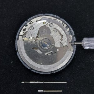 May include: A close-up view of a watch movement with intricate gears and components. The watch mechanism is silver and gray, with the text "NH35A" and "TWENTY-FOUR JEWELS" visible. A small screwdriver is inserted into the mechanism.