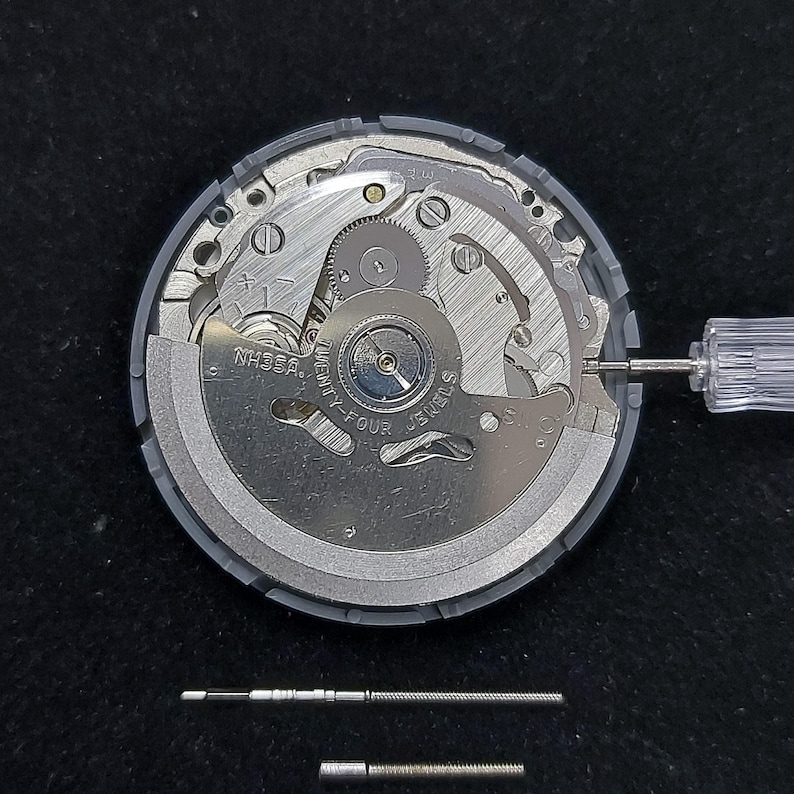 May include: A close-up view of a watch movement with intricate gears and components. The watch mechanism is silver and gray, with the text "NH35A" and "TWENTY-FOUR JEWELS" visible. A small screwdriver is inserted into the mechanism.