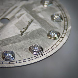 May include: Close-up of a watch face with a textured, gray surface and the word "Automatic" in cursive. The face features several square-cut, sparkling gemstones set around the perimeter, adding a touch of luxury to the design.