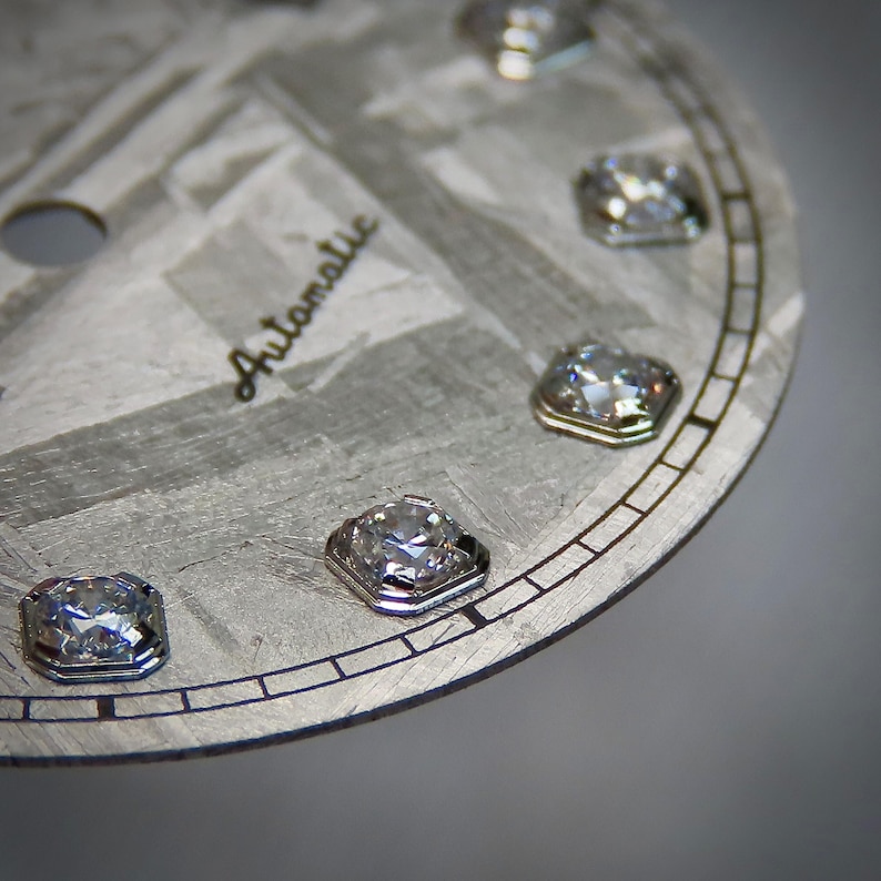 May include: Close-up of a watch face with a textured, gray surface and the word "Automatic" in cursive. The face features several square-cut, sparkling gemstones set around the perimeter, adding a touch of luxury to the design.