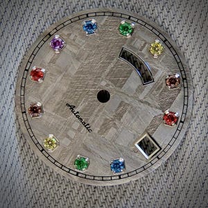 May include: A watch face with a textured, gray surface and a black border. It features twelve colorful gemstones and a black dial. The word "Automatic" is printed in a script font. The watch face has a unique, patterned design.