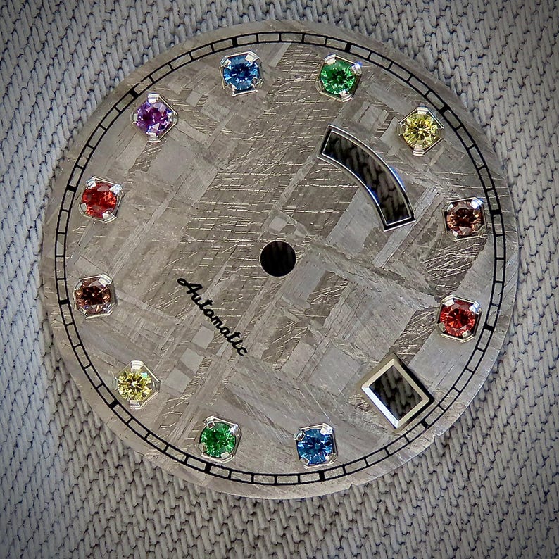 May include: A watch face with a textured, gray surface and a black border. It features twelve colorful gemstones and a black dial. The word "Automatic" is printed in a script font. The watch face has a unique, patterned design.