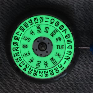 Puede incluir: Un disco de calendario circular para movimiento de reloj con fondo verde luminoso y texto impreso en negro. El disco muestra los números del 1 al 31 y los días de la semana en inglés y japonés, dispuestos en un patrón radial.