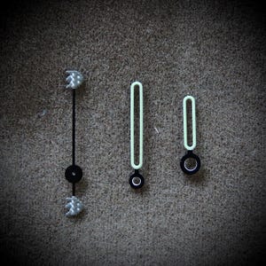 May include: Close-up of watch hands. The set includes a black second hand with textured silver accents, and two luminous green hands with black bases. The hands are designed for a watch.