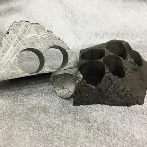 May include: Two pieces of textured, gray stone. One is triangular with two circular cutouts. The other is a dark, irregular shape with multiple openings. The stone has a unique, cross-hatched pattern.