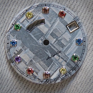May include: A watch face with a textured gray background and a grid-like pattern. It features a silver-colored curved element, a small square, and the word "Automatic". The face is adorned with colorful gemstones.