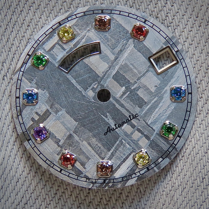 May include: A watch face with a textured gray background and a grid-like pattern. It features a silver-colored curved element, a small square, and the word "Automatic". The face is adorned with colorful gemstones.