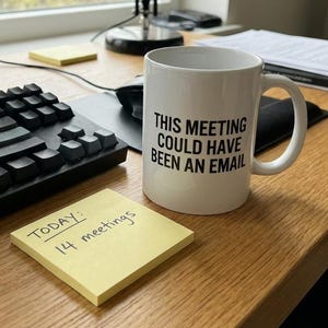 Puede incluir: Una taza de cerámica blanca con el texto "THIS MEETING COULD HAVE BEEN AN EMAIL" en letras negras. Una nota adhesiva amarilla dice "TODAY: 14 meetings". La taza está sobre un escritorio de madera junto a un teclado.