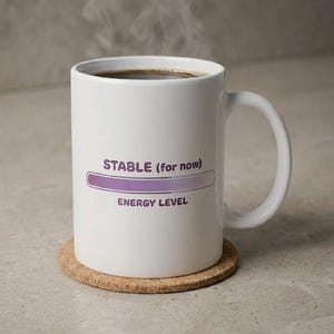 Puede incluir: Una taza de cerámica blanca con asa, llena de un líquido oscuro que desprende vapor. El frente presenta texto morado que dice STABLE (for now) sobre una barra de progreso morada y el texto ENERGY LEVEL impreso debajo de la barra en morado.