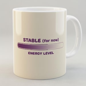 Puede incluir: Una taza de cerámica color crema con un asa blanca. La taza presenta el texto "STABLE (for now)" encima de una barra de nivel de energía púrpura, y las palabras "ENERGY LEVEL" debajo. La taza está diseñada para bebidas.