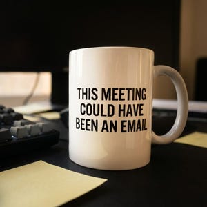 May include: A white ceramic mug with a handle, featuring the black printed text THIS MEETING COULD HAVE BEEN AN EMAIL. The mug has a cylindrical shape and a smooth finish, standing upright as a functional beverage container for hot or cold drinks.