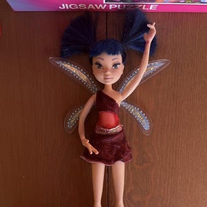 Könnte beinhalten: Eine Feenpuppe mit dunkelblauem Haar in zwei Zöpfen, bekleidet mit einem dunkelroten, asymmetrischen Kleid mit Taillenausschnitt. Die Puppe hat durchscheinende Glitzerflügel und steht auf einem roten Kunststoffständer. Der Text JIGSAW PUZZLE ist sichtbar.