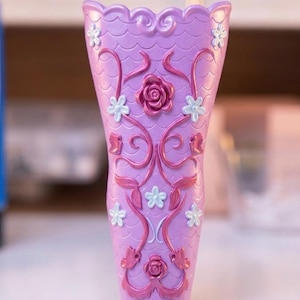 Può includere: Un vaso color lavanda con bordo smerlato e un motivo a scaglie strutturato sulla superficie. L'esterno è decorato con motivi di rose metalliche rosa in rilievo, dettagli a viticcio vorticosi e piccoli accenti floreali azzurri a cinque petali.