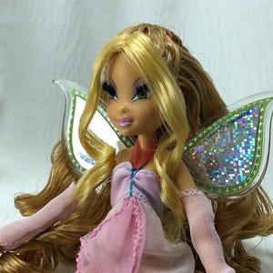 May include: A doll with long blonde hair, green eyes, and purple eyeshadow. The doll wears a light pink sleeveless dress with a red strap and sheer pink arm sleeves. Large, translucent, iridescent wings with green borders are attached to the back.