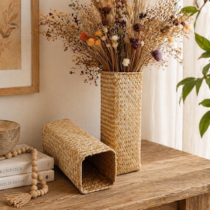 Könnte beinhalten: Zwei rechteckige gewebte Vasen, eine aufrecht und gefüllt mit getrockneten Blumen in Braun-, Orange- und Lilatönen. Die andere Vase liegt auf der Seite. Beide Vasen bestehen aus einem hellbraunen gewebten Material und stehen auf einer Holzoberfläche.