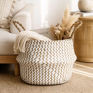 Puede incluir: Una cesta tejida con un patrón de chevrón blanco y beige. La cesta tiene dos asas de cuerda y está llena de flores secas y una manta color crema. La cesta está sobre una alfombra beige.