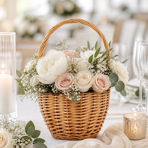 May include: A round wicker basket brimming with white peonies, pale pink roses, and foliage. The basket has a curved handle and rests on a white tablecloth. A lit candle in a glass holder and a white runner are also present.
