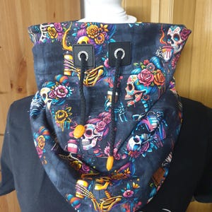 Könnte beinhalten: Ein schwarzer Cowl-Neck-Wärmer mit einem farbenfrohen Tag der Toten Schädel- und Blumendruck. Der Nackenwärmer hat schwarze Kordelzüge mit orangefarbenen Knebeln und schwarzen Kunstlederakzenten. Das Design zeigt Totenköpfe, Blumen und Skelette.