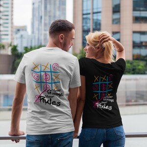 May include: Two people wearing matching t-shirts with a tic-tac-toe design on the back. One shirt is white, the other is black. The design includes a grid, smiley faces, and the text "Your Game Your Rules" in a graffiti style.