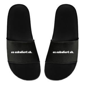 May include: A pair of black slide sandals. Each sandal has a wide strap with the phrase "es eskaliert eh." printed in white. The sandals are designed for casual wear.