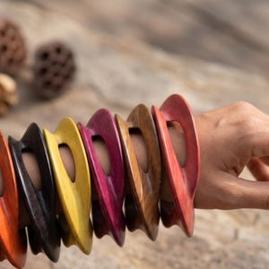 Peut inclure: Un ensemble de sept bracelets rigides en bois de différentes couleurs, notamment bleu sarcelle, orange, noir, jaune, magenta, marron et rouge. Chaque bracelet présente un design incurvé ajouré avec une finition lisse, portés empilés.