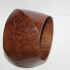 Pode incluir: Uma pulseira de madeira larga e de paredes grossas com um acabamento polido em castanho avermelhado escuro. A pulseira apresenta uma superfície exterior lisa e arredondada com padrões de grão de madeira natural visíveis e uma abertura circular.