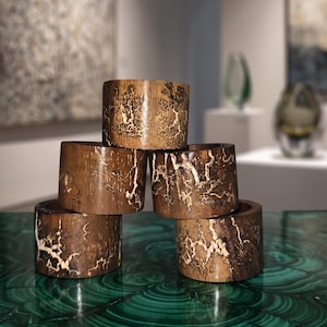 May include: A stack of five cylindrical wooden napkin rings featuring a dark brown finish with intricate, light-colored branching patterns across the surface. The rings are arranged in a pyramid formation, showcasing the natural wood grain and texture.