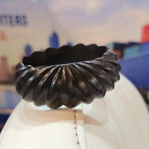 Könnte beinhalten: Ein dunkelbrauner, kreisförmiger Armreif mit einer tief geschnitzten, gewellten oder geriffelten Textur auf der gesamten Außenfläche. Der Armreif hat eine glatte, polierte Oberfläche und eine gleichmäßige, abgerundete Form entlang seines gesamten Umfangs.