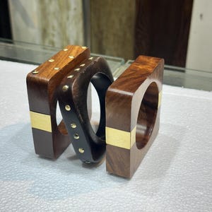 May include: Three square-shaped wooden bangles. Two bangles are made of polished brown wood with a smooth gold-tone metal band inlay. The central bangle is dark brown wood featuring a row of small, round, gold-tone metal studs along the outer edge.