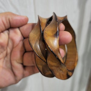 Puede incluir: Dos brazaletes circulares de madera con una silueta retorcida y ondulada. Las pulseras presentan un acabado pulido con patrones de vetas de madera natural en varios tonos de marrón medio a oscuro, resaltando los bordes curvos.