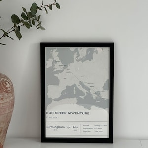 May include: A framed print featuring a map of Europe with a flight path from Birmingham to Kos, Greece. The text "OUR GREEK ADVENTURE" is displayed, along with flight details. The frame is black, and the print is in grayscale.