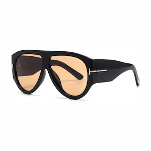 May include: Oversized sunglasses featuring a thick black frame and a continuous brow bar design. The lenses are a light amber or peach color. A silver-toned T-shaped metal accent is visible on the temple hinge, connecting the frame to the arm.