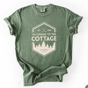 May include: Moss green t-shirt with a white graphic. The graphic reads "I'M COMING TO THE COTTAGE" inside a white hexagon with the text "EST 2025" and a tree line. The word "CANADA" is below the tree line.