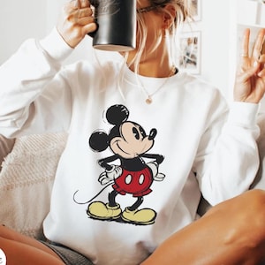 Pode incluir: Camisola branca com um gr&aacute;fico do Mickey Mouse em preto, vermelho e amarelo. A camisola tem gola redonda e mangas compridas. A palavra "White" est&aacute; num c&iacute;rculo branco no canto inferior esquerdo. O design &eacute; uma imagem cl&aacute;ssica de desenho animado.