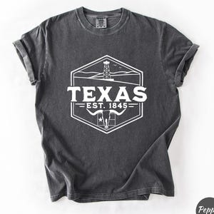 May include: Dark grey t-shirt featuring a white graphic of the state of Texas. The graphic includes the word "TEXAS" above "EST. 1845" and a depiction of a Texas longhorn, oil derrick, and a star. The shirt has rolled-up sleeves.