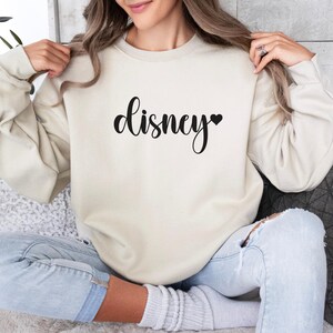 May include: Cream-colored sweatshirt with the word "Disney" in a black script font, a black heart replaces the letter "y". The long-sleeved top is made of a soft material.