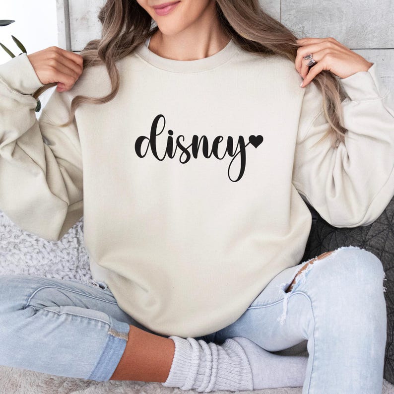 May include: Cream-colored sweatshirt with the word "Disney" in a black script font, a black heart replaces the letter "y". The long-sleeved top is made of a soft material.