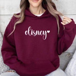 May include: Burgundy hoodie with a front pocket and a white cursive "disney" text with a heart symbol. The hoodie is made of a soft material and has a hood.
