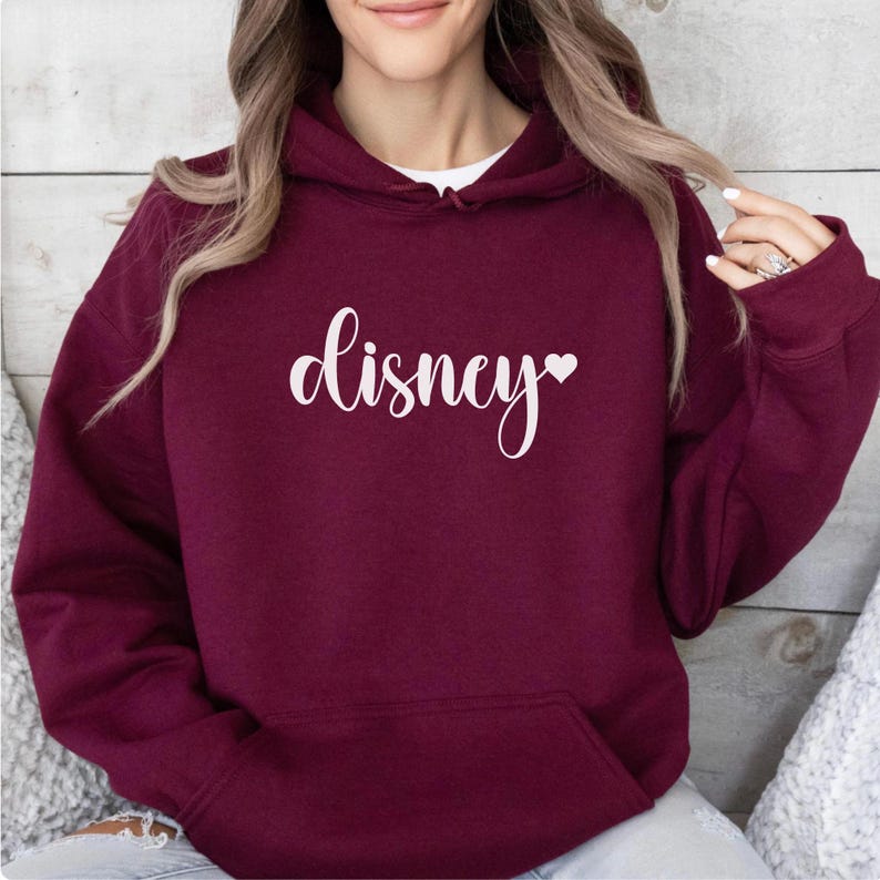 May include: Burgundy hoodie with a front pocket and a white cursive "disney" text with a heart symbol. The hoodie is made of a soft material and has a hood.