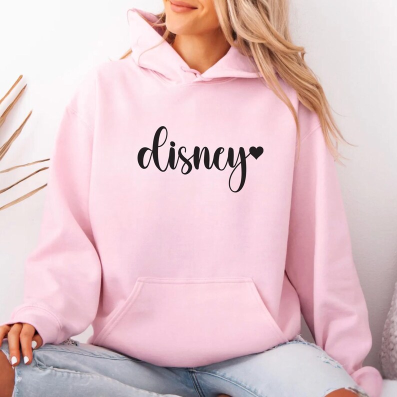 May include: A light pink hoodie with a front pocket and a hood. The word "Disney" is printed in black script across the chest, with a small black heart replacing the letter "y".