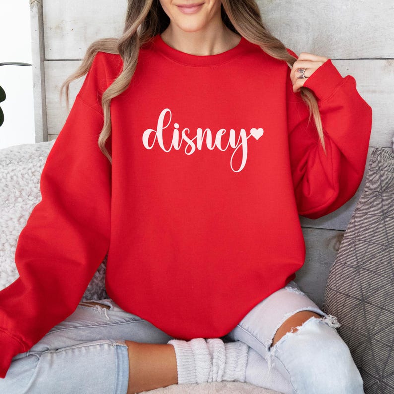 May include: A red sweatshirt with the word "Disney" in white script, with a small heart symbol. The sweatshirt is a solid color and has a crew neck. The sleeves are long and the material appears to be soft.