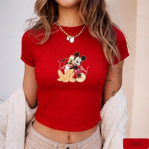Puede incluir: Un top corto rojo con un gr&aacute;fico de Mickey Mouse y Pluto abraz&aacute;ndose. Los personajes de dibujos animados est&aacute;n representados en un abrazo amistoso, con Mickey con su atuendo ic&oacute;nico y Pluto sentado. La parte superior es de color rojo brillante.