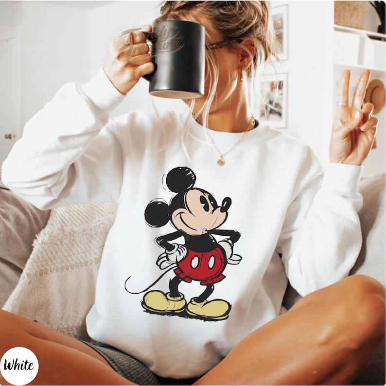 Pode incluir: Camisola branca com um gr&aacute;fico do Mickey Mouse em preto, vermelho e amarelo. A camisola tem gola redonda e mangas compridas. A palavra "White" est&aacute; num c&iacute;rculo branco no canto inferior esquerdo. O design &eacute; uma imagem cl&aacute;ssica de desenho animado.
