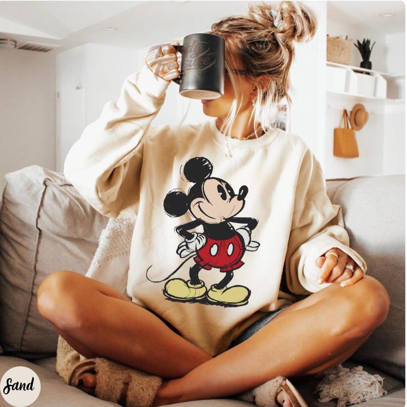 Pode incluir: Camisola creme com um grande gr&aacute;fico do Mickey Mouse. Mickey &eacute; retratado com contornos pretos, cal&ccedil;&otilde;es vermelhos, sapatos amarelos e luvas brancas. A camisola tem gola redonda e mangas compridas. Uma caneca preta &eacute; segurada perto do rosto.